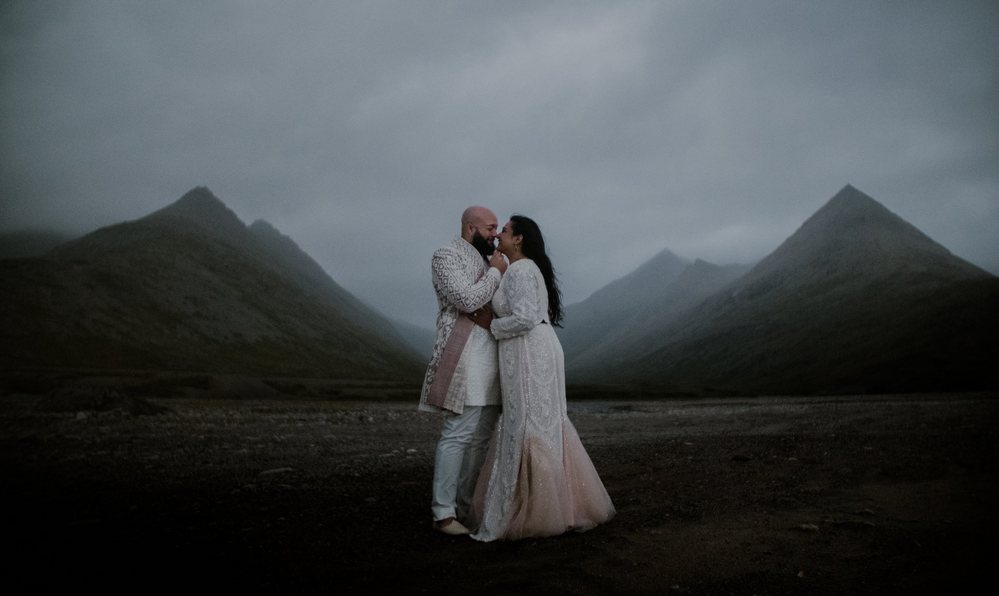 A couple holds each other close on black sand before the sharp peaks of Stokksnes in Iceland under a heavy grey sky.