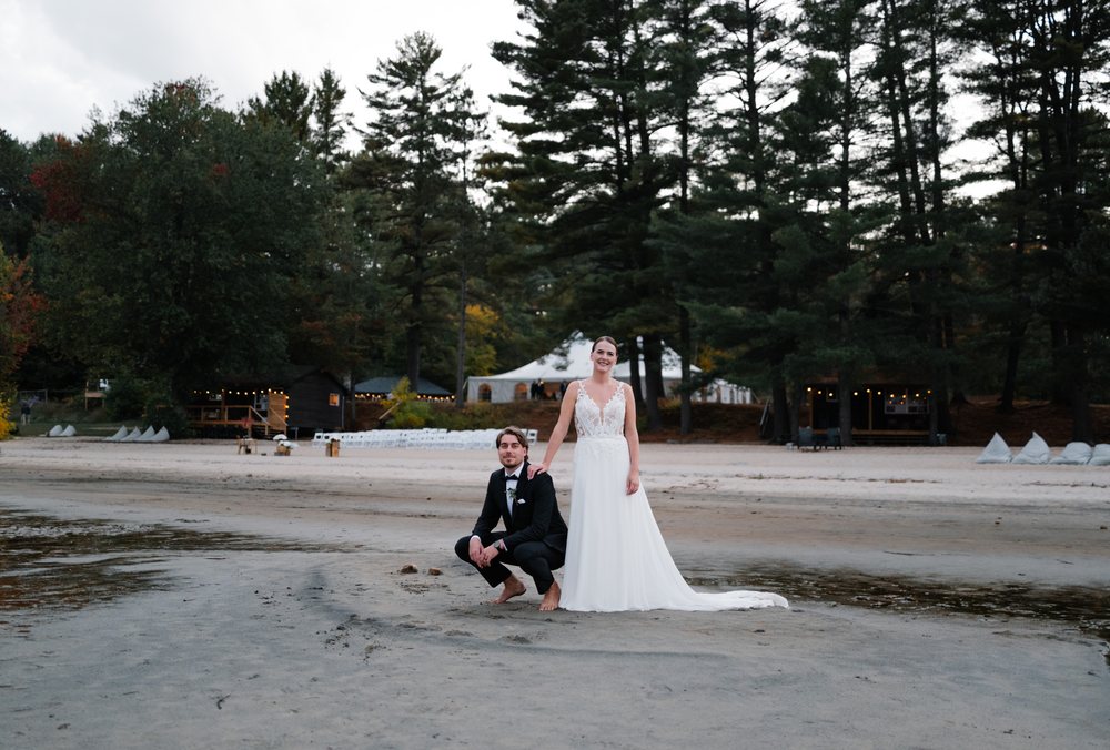 A bride stands barefoot on the sand at Anupaya Cabin Co. while her groom crouches beside her holding her hand. Cabins and tall pines behind them.
