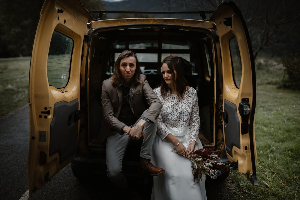 A couple in wedding clothes sits side by side in the open back of a yellow van on a country road. The bride holds a bouquet of dried flowers.