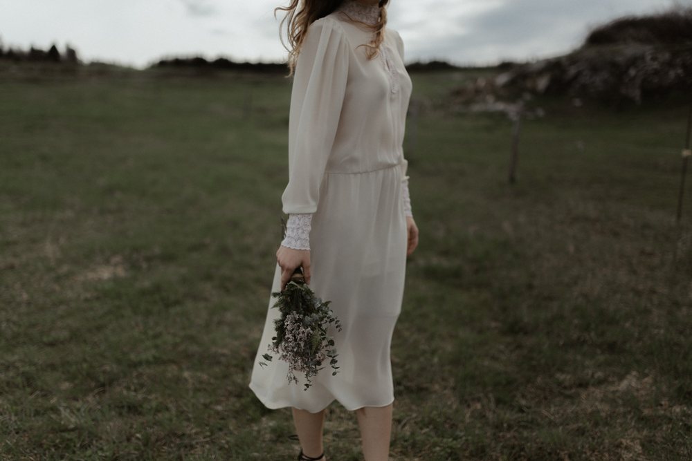 A bride in a long ivory dress stands in a grassy field and holds a small bouquet down at her side.