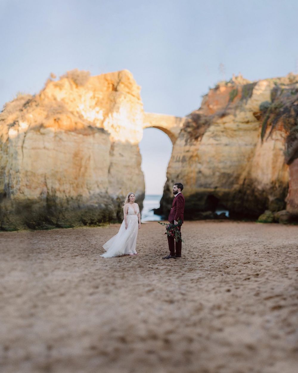 A golden rock arch on a beach in the Algarve, a couple standing beneath it by the sea.