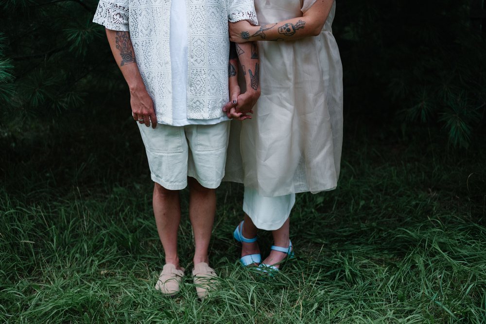 A couple in white summer clothes holds hands in tall green grass among the pines.