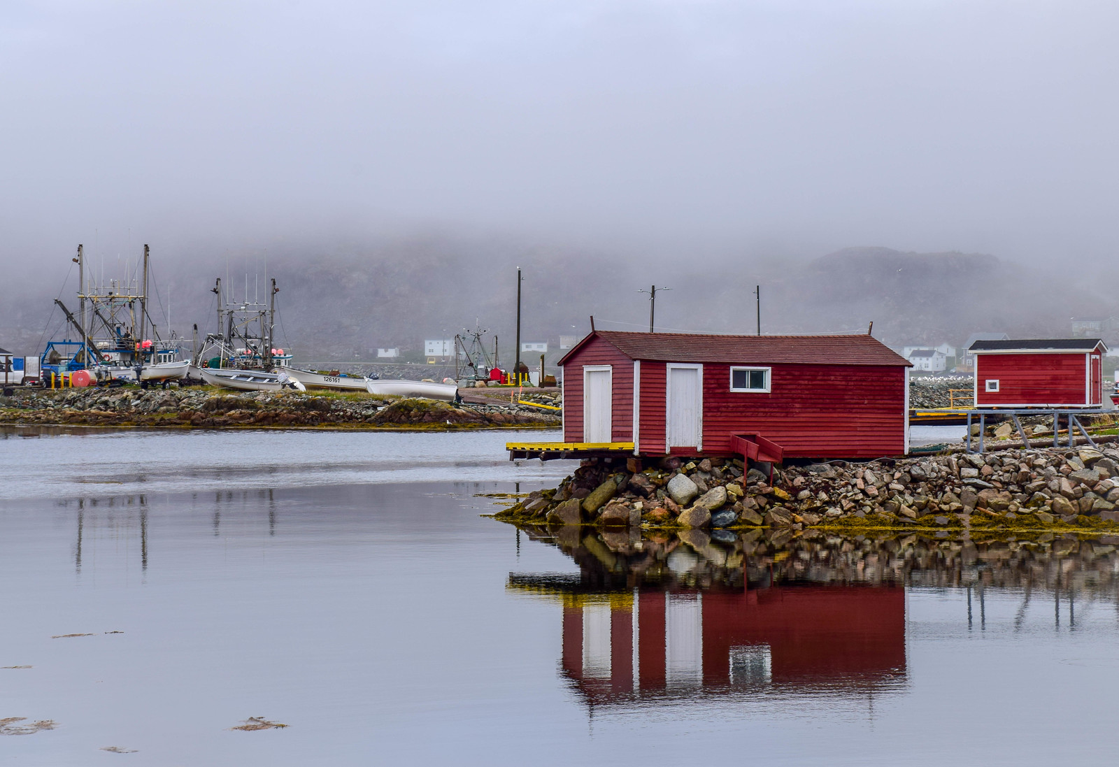 A red fishing stage reflected in the still harbour water of Fogo Island, Newfoundland, under low fog
