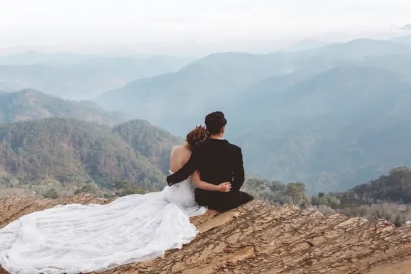 A couple seen from behind on a rock ridge, her dress spread over the stone, layered blue mountain ranges fading into the mist