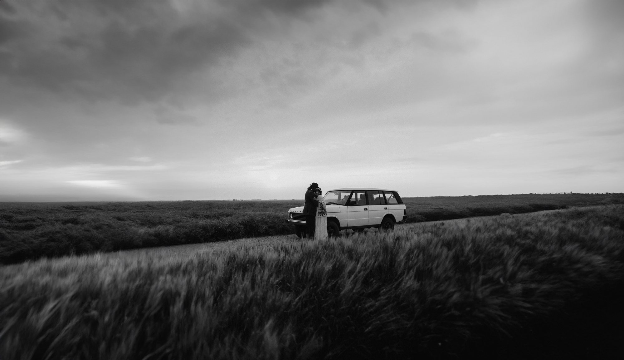 A couple holds each other beside a white station wagon on a gravel road crossing the Saskatchewan prairie. Tall grass in front of them and a storm sky over everything. Black and white.